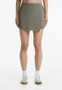 Jupe portefeuille vert olive au tissu lisse, dotée d'une taille élastique et d'un détail de nœud sur le côté. Associée à des baskets claires et des chaussettes blanches.
