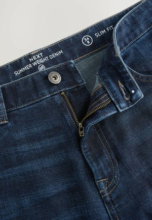 Jeans in denim slim fit di colore blu scuro con zip, bottone in metallo, cuciture a contrasto e un'etichetta che indica "Denim Estivo".