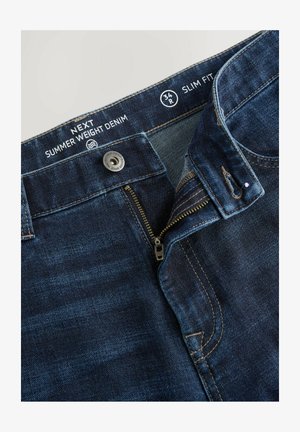 Donkerblauwe slim fit denim jeans met een rits, metalen knoop, contrasterende stiksels en een label met de tekst "Zomergewicht Denim."