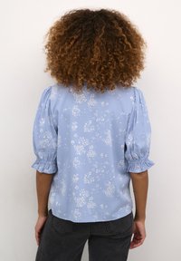Lichtblauwe blouse met korte, opgezette mouwen, voorzien van een witte bloemmotief en elastische manchetjes. De stof lijkt zacht met een gladde textuur.