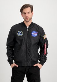 Black bomber jacket with NASA patches, ribbed cuffs, and a zipper. Features a red tag on the sleeve with text, smooth fabric, and front pockets.