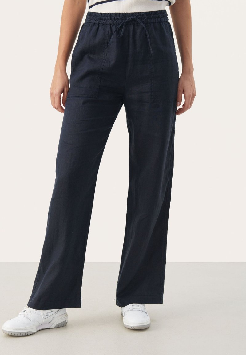 Femme portant un pantalon ample bleu marine foncé avec une ceinture élastique et des baskets blanches, debout sur un sol clair.
