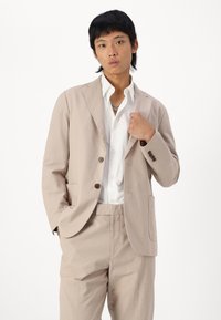 Beige textured suit with matching pants and a white shirt. Features a two-button closure, notch lapels, and side pockets.