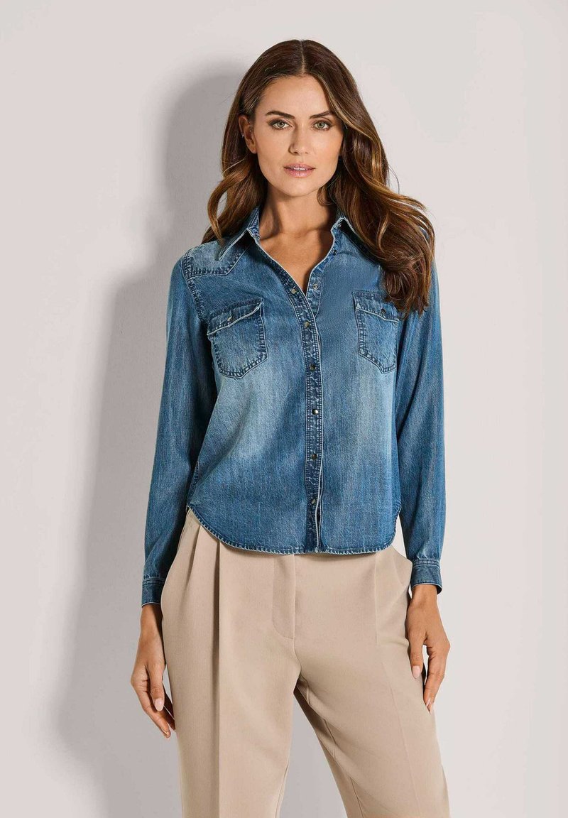 Lichtblauw denim overhemd met lange mouwen, knoopsluiting aan de voorkant, twee borstzakken en een gebogen zoom; gecombineerd met beige plooibroek.