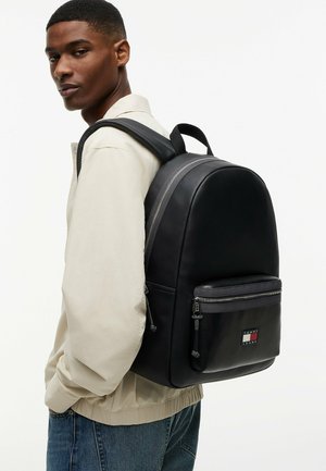 Giovane uomo con giacca beige e jeans blu porta uno zaino nero Tommy Jeans con tasca frontale e cerniere argentate.