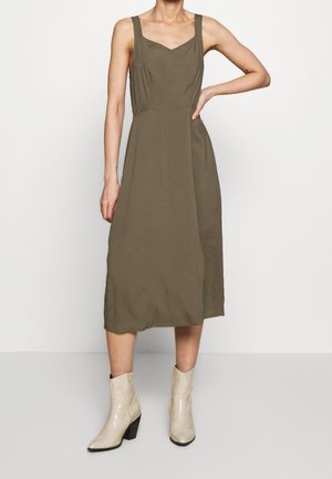 Olivengroene jurk met een getailleerd lijfje, vierkante halslijn en midilange rok. Draagt lichte, textuurrijke enkellaarsjes.