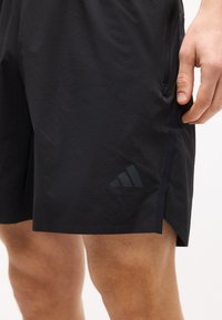 Μαύρα αθλητικά σορτς σε άτομο, με διακριτικό λογότυπο Adidas κοντά στο τελείωμα και τσέπη με φερμουάρ στο πλάι.