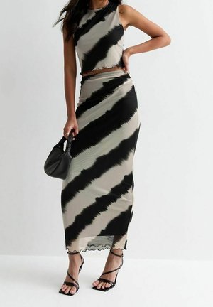 Frau trägt schwarz-beiges batikartiges, ärmelloses Crop-Top und passenden langen Rock, hält kleine schwarze Tasche, trägt schwarze Riemchen-High Heels.