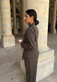 Hnedý formálny oblek s vysokým golierom a štruktúrovanými ramenami. Tkanina vyzerá hladko. Drží kávový hrnček a stojí pri kamenných stĺpoch.