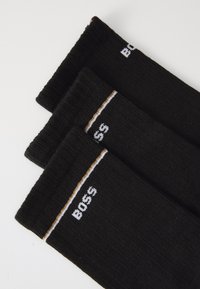 Μαύρες κάλτσες με ριγέ υφή, που διαθέτουν λευκό λογότυπο "BOSS" και μία οριζόντια χρυσή ρίγα στον ποδόγυρο. Τρία ζευγάρια στοιβαγμένα.