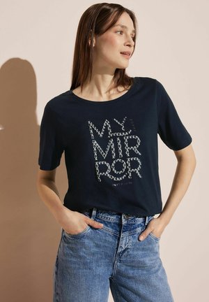 T-shirt in cotone blu navy con maniche corte, dotata di una stampa grafica testurizzata in bianco e argento che recita "IL MIO SPECCHIO." Abbinata con jeans blu.