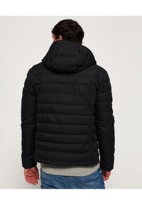 Sort polstret jakke med hætte, der har et kviltet design og tekstureret stof. Synlig grå sweater i bunden og blå jeans.