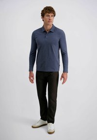 Mann steht da in einem blauen Langarm-Polohemd, schwarzen Hosen und weißen Sneakers vor einem schlichten hellen Hintergrund.