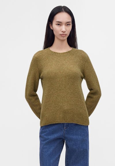 Femme aux longs cheveux noirs portant un pull en tricot vert olive et un jean bleu, debout les mains derrière le dos sur un fond uni.
