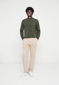 Pull en laine noir avec des manches longues et un col rond, associé à un pantalon beige clair et des baskets blanches. Logo jaune sur le pull.