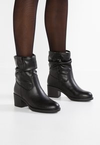 Bottines en cuir noir avec un design décontracté, détail de fermeture éclair sur le côté, talon empilé et texture lisse. Portées avec des collants noirs transparents.
