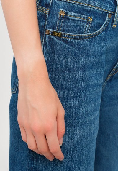 Jean en denim bleu avec des poches visibles, des coutures dorées et une petite étiquette noire portant la mention "tiger of sweden." Gros plan avec une main posée.