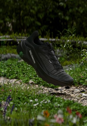 Chaussure de running Salomon noire avec un design en mesh et des logos de la marque, montrée au-dessus d'un chemin de jardin rocailleux et fleuri avec un feuillage vert en arrière-plan.