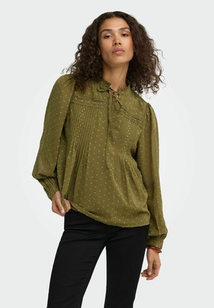 Femme aux cheveux bouclés portant un chemisier vert olive à manches longues avec des détails en dentelle et des plis, associé à un pantalon noir sur fond blanc.