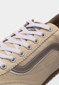 Gros plan d'une basket en daim beige avec des lacets blancs passés dans des œillets métalliques et une couture ondulée marron sur la bande latérale.
