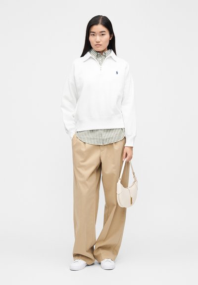 Sudadera blanca con cremallera y cuello, superpuesta a una camisa de rayas verdes y blancas. Pantalones beige de corte holgado y un bolso pequeño beige.