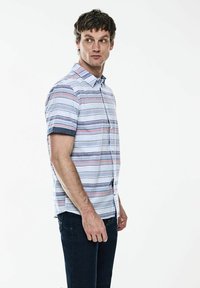 Košile s krátkým rukávem v světle modré barvě s horizontálními barevnými pruhy. Má klasický límec a námořnické akcenty na manžetách.