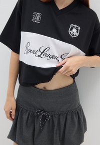 Jersey deportivo en blanco y negro con cuello en V, que presenta una textura de malla, un logotipo y la impresión "Sport League". Combinado con una falda gris con volantes.