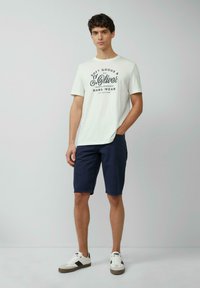 Męski jasnoszary t-shirt z czarnym tekstem graficznym, zestawiony z granatowymi szortami i białymi trampkami z czarnymi i szarymi akcentami.