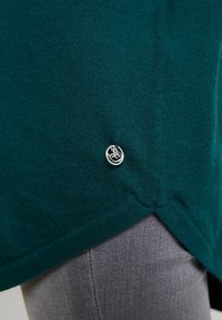 Türkisfarbener, gestrickter Pullover mit lockerem Schnitt, ausgestattet mit einem silbernen Logoakzent am Saum und einem geschwungenen, geteilten Design am unteren Rand.