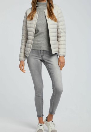 Femme portant une veste matelassée beige clair sur un pull à col roulé gris, un jean skinny gris et des baskets blanches, debout sur un fond uni.