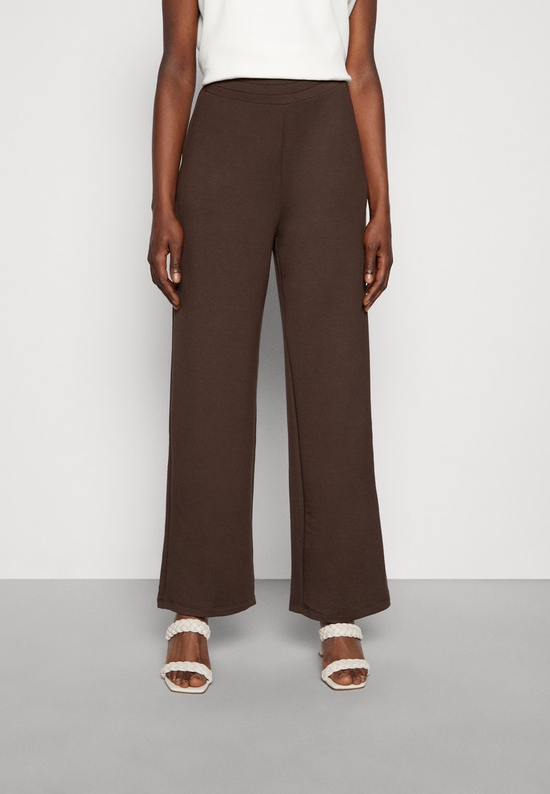 Pantalons larges marron avec une taille élastique, fabriqués en tissu doux. Portés avec des sandales blanches à brides tressées, mettant en valeur une texture lisse.