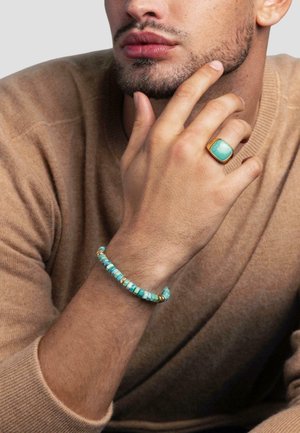 Turquoise ring with gold accents worn on a hand, alongside a beaded bracelet of alternating turquoise blue and gold beads. Worn with a beige sweater.