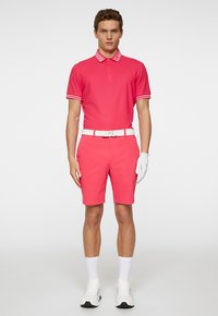 Herrpolo i rosa med vit krage och ärmar, kombinerat med rosa shorts, vit bälte och vita träningsskor. Handske synlig.