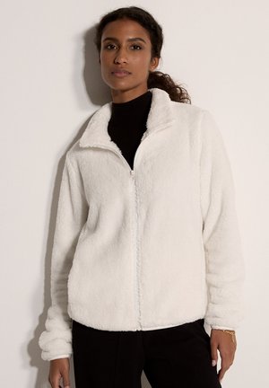 Frau trägt eine weiße, flauschige Jacke mit Reißverschluss über einem schwarzen Oberteil, steht vor einem einfarbigen hellen Hintergrund mit neutralem Gesichtsausdruck.