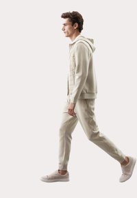 Beige zip-up hoodie over en lys beige langærmet skjorte, parret med matchende tapered bukser og lys beige sneakers, gående i profil.