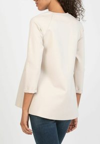 Light beige three-quarter sleeve top with rounded hem and rolled cuffs, made of a smooth, textured fabric. Back view shows minimal detailing.