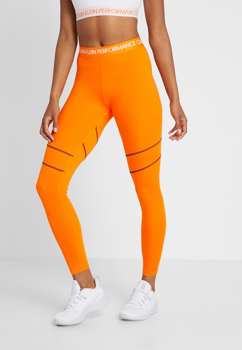 Jasnopomarańczowe legginsy sportowe z wysokim stanem i kontrastującymi czarnymi szwami, noszone z białymi butami sportowymi.