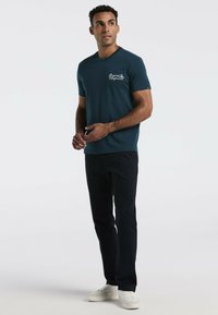 Teal crew-neck t-shirt met korte mouwen en witte logodetail, gecombineerd met zwarte broek en witte sneakers. Soepele stoftextuur.