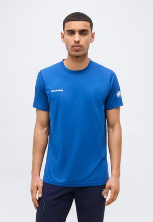 Άνδρας που φοράει μπλε κοντομάνικο πουκάμισο Mammut και μαύρο παντελόνι, στέκεται μπροστά σε ένα απλό ανοιχτόχρωμο φόντο.