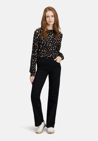 Jonge vrouw met lang rood haar draagt een zwart-wit gestippelde blouse met lange mouwen, zwarte jeans en witte sneakers, staand met haar hand in haar zak.