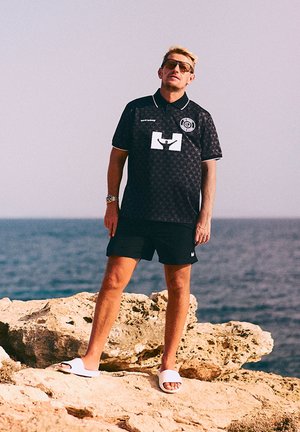 Schwarzes Polohemd mit einem sich wiederholenden geometrischen Muster, weißem Logo-Design, kombiniert mit schwarzen Shorts und weißen Slide-Sandalen. Küstenkulisse.
