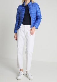 Blå puffjacka med dragkedja framtill, rund krage och elastiska ärmar, i kombination med vita raka jeans och vita sneakers.