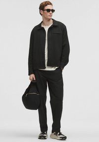 Giacca nera leggera sopra una maglietta bianca, abbinata a pantaloni neri. Gli accessori includono occhiali da sole scuri e una borsa nera con cerniera dorata.