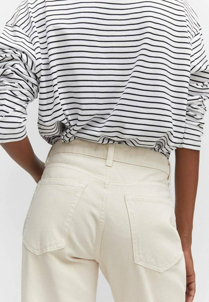 Langärmliges, gestreiftes weißes Hemd, das mit hochtaillierten cremefarbenen Jeans getragen wird. Die Jeans verfügen über Gesäßtaschen und eine genähte Taillenlinie.