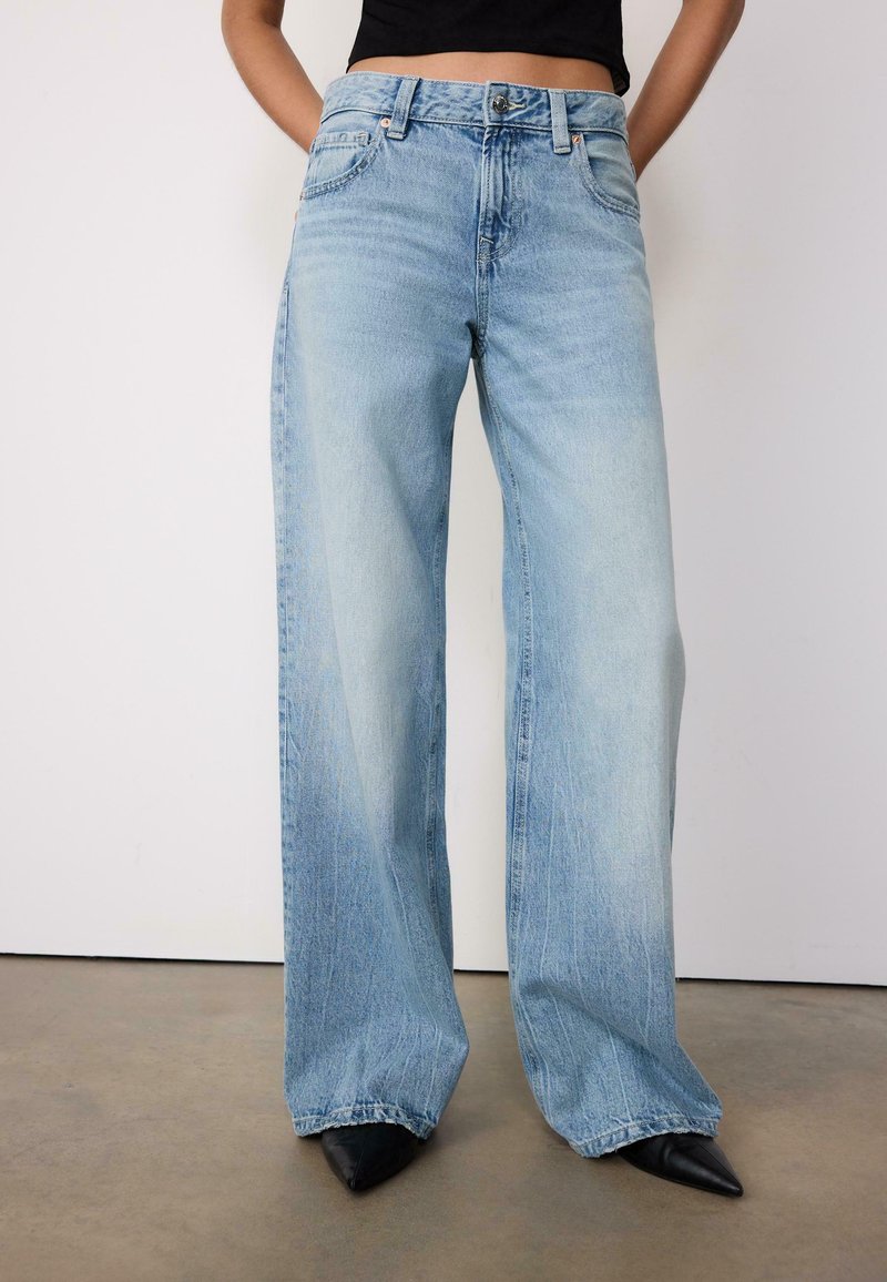 Personne portant un jean taille haute bleu clair à jambes larges et des chaussures noires pointues, debout sur un sol en béton contre un mur blanc.