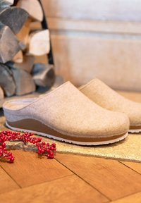 Beige Filzpantoletten mit geschwungener Form, weißer Gummisohle und braunem Akzent. Rote dekorative Beeren in der Nähe auf einer strukturierten Oberfläche.