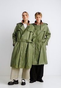 Δύο άτομα φορούν μακριά, χακί χακί παλτό με ζώνες και μεγάλα κουμπιά. Το ένα έχει ένα καφέ γιακά, ενώ και τα δύο φορούν στρωμένα ρούχα με παντελόνια με μοτίβα.