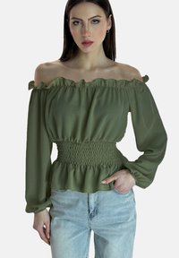 Olivgrüne Off-Shoulder-Bluse mit Rüschenausschnitt und smockiertem Taillenbund, kombiniert mit hellblauen Jeans, langen Ärmeln und einem Peplumsaum.