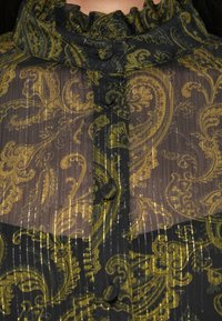 Črna prosojna bluza z vzorcem paisley v zlati in zeleni barvi, z volančnim ovratnikom in gumbi. Teksturirana z navpičnimi črtami.