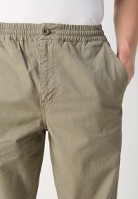 Leichte, olivgrüne Hose mit einem elastischen Bund, Fronttaschen und einem Knopfverschluss. Glattes Material mit einer leicht zerknitterten Textur.
